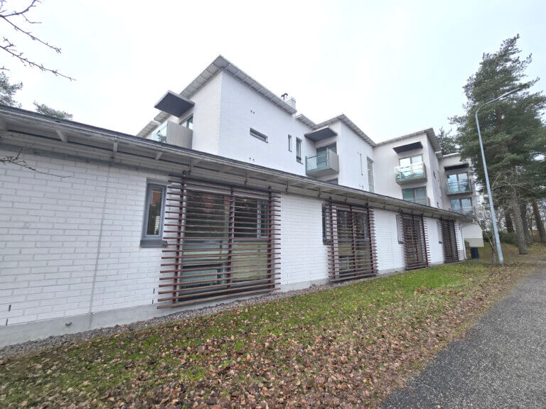 Moderni, vaaleaksi maalattu asuinrakennus, jossa on lasitetut parvekkeet ja puiset sälekaihtimet, sijaitsee puistomaisessa ympäristössä.