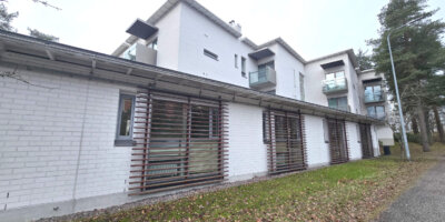 Moderni, vaaleaksi maalattu asuinrakennus, jossa on lasitetut parvekkeet ja puiset sälekaihtimet, sijaitsee puistomaisessa ympäristössä.