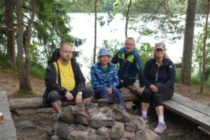 Neljä ihmistä kesäisessä luonnossa nuotiopaikan ympärillä taustalla järvi
