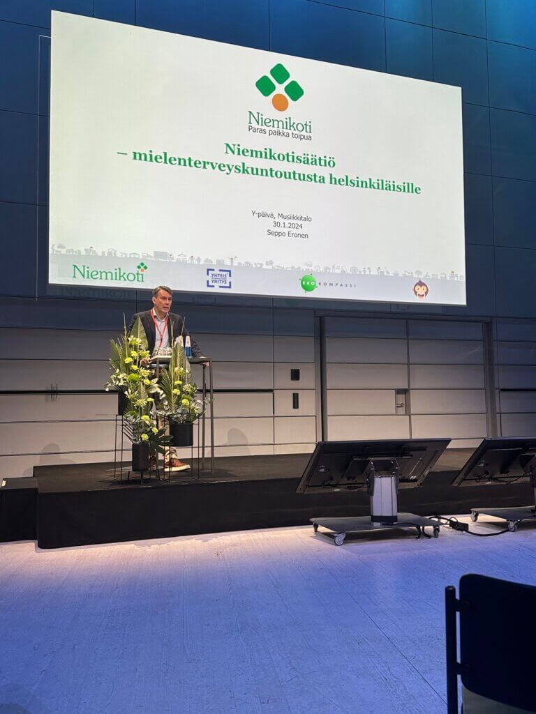 Kuva esiintymislavasta, jossa Seppo Eronen puhujapöntössä taustalla powerpoint-esitys.