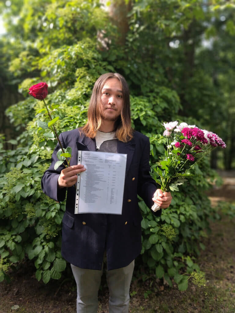 Nuori henkilö seisoo ulkona vehreässä ympäristössä pitäen isoa paperiarkkia edessään. Hänellä on toisessa kädessä punainen ruusu ja toisessa kädessä kimppu värikkäitä kukkia.