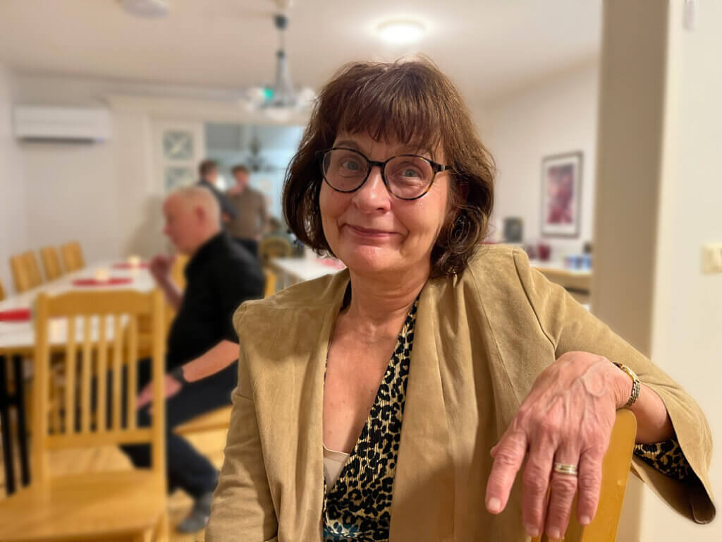 Nainen hymyilee kameraan sisätilassa. Hänellä on lasit ja beige takki. Taustalla näkyy muutamia ihmisiä ja ruokapöytiä.