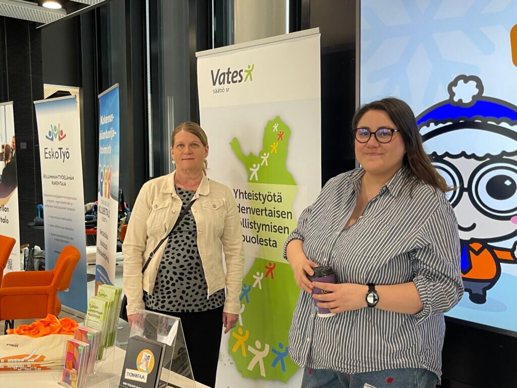 Kaksi henkilöä seisoo messupöydän edessä sisätiloissa. Taustalla näkyy useita julisteita ja mainoskylttejä, joissa on erilaisia logoja ja tekstejä, kuten Vates-säätiö ja EskoTyö. Oikealla on myös värikäs piir
