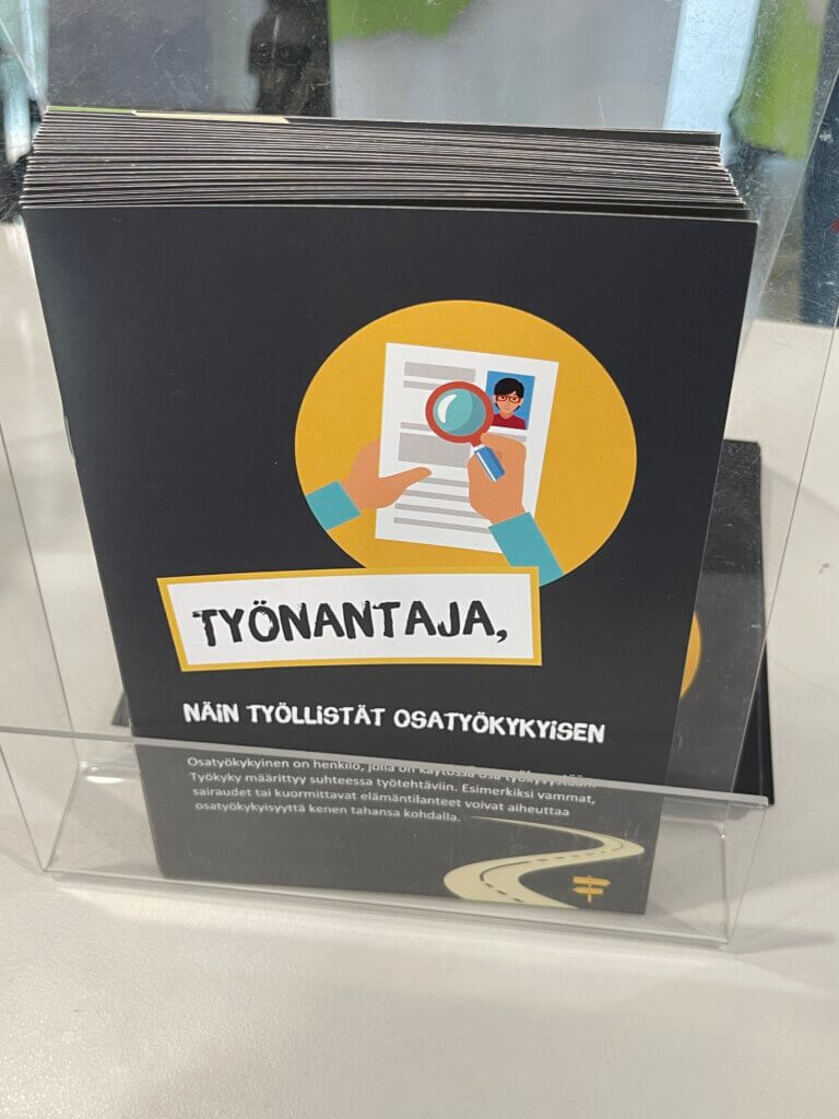 Esitteitä telineessä. Päällimmäisessä esitteessä on kädet, jotka pitelevät suurennuslasia henkilön kuvaa ja tekstiä sisältävän paperin päällä. Esite on suunnattu työnantajille ja siinä lukee: Työnantaja, näin työllistät os