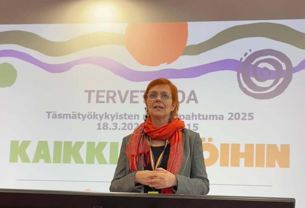 Nainen, jolla on punainen huivi, puhuu lavalla tapahtumassa. Hänen takanaan näkyy tervetulotoivotus ja tapahtuman nimi Täsmätyökykyisten päivä - tapahtuma 2025.