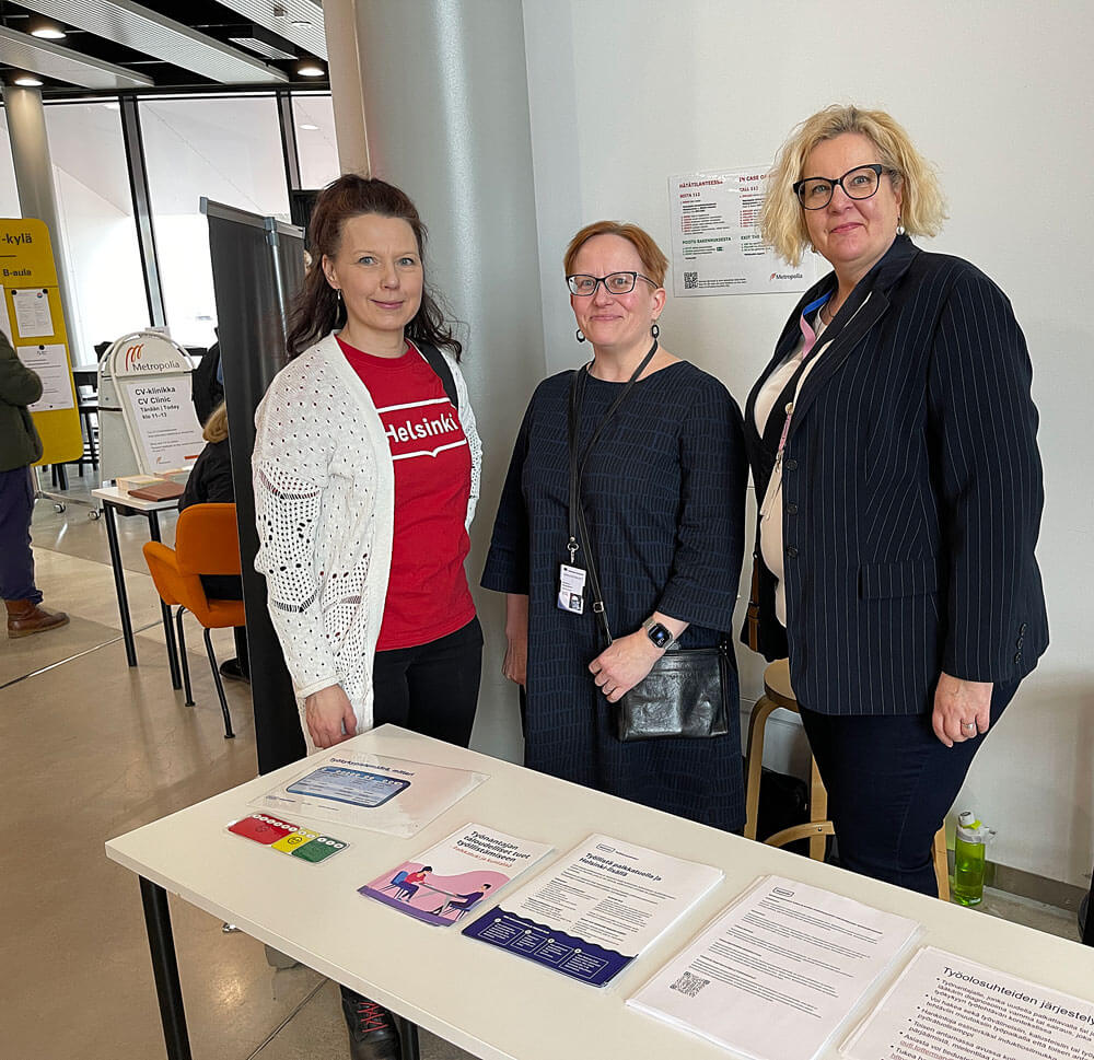 Kolme henkilöä seisoo pöydän ääressä sisätiloissa. Pöydällä on erilaisia esitteitä ja asiakirjoja. Vasemmalla henkilö, jolla on punainen Helsinki -t-paita ja valkoinen neule. Keskellä ja oikealla olevat henkilöt ovat puke
