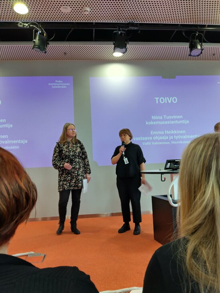 Kuvassa kaksi henkilöä seisoo esityksessä lavalla. Heillä on mikrofonit ja he puhuvat yleisön edessä. Taustalla näkyy projektiokangas, jossa on tekstiä liittyen esitykseen. Osa yleisöstä on etualalla.