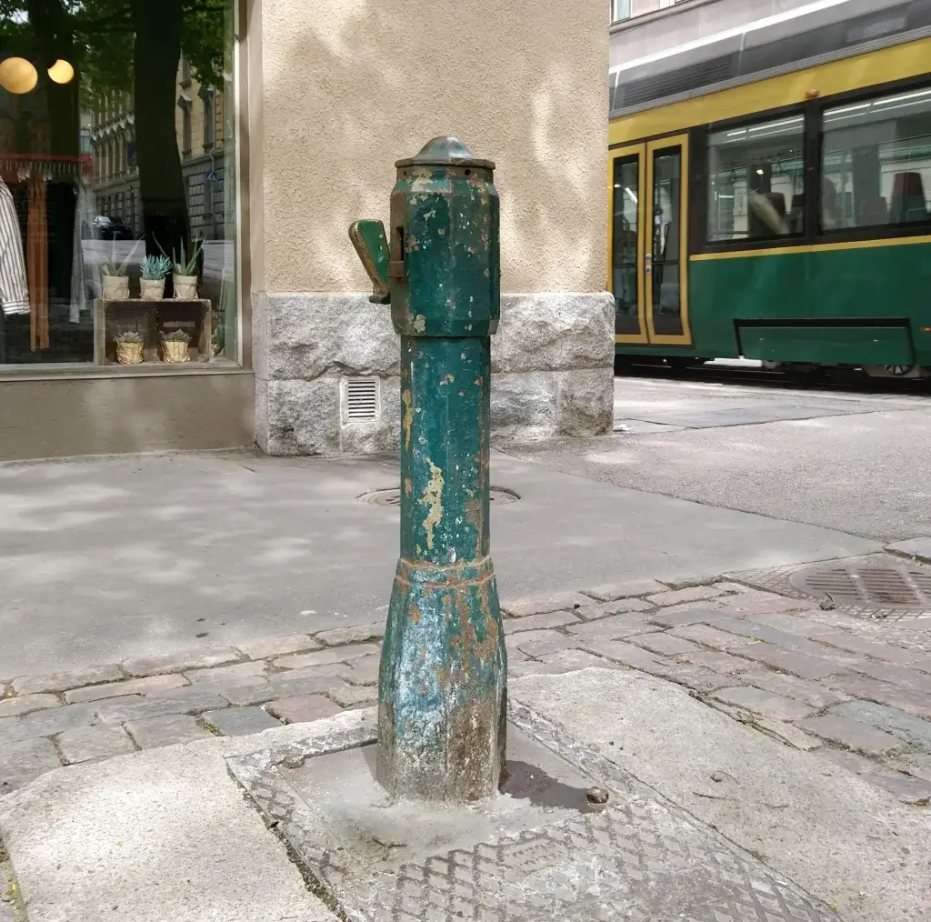 Vanhanaikainen vihreä vesiposti kadulla, jossa raitiovaunu kulkee ohi taustalla. Vasemmalla näkyy kaupan ikkuna, jossa on sisustuskasveja.