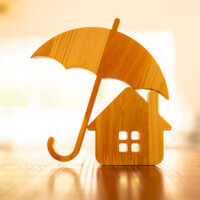 Wooden models depicting a house and an umbrella are placed on a wooden surface, with the umbrella positioned to shelter the house. The background is softly blurred, creating a warm, inviting ambiance.