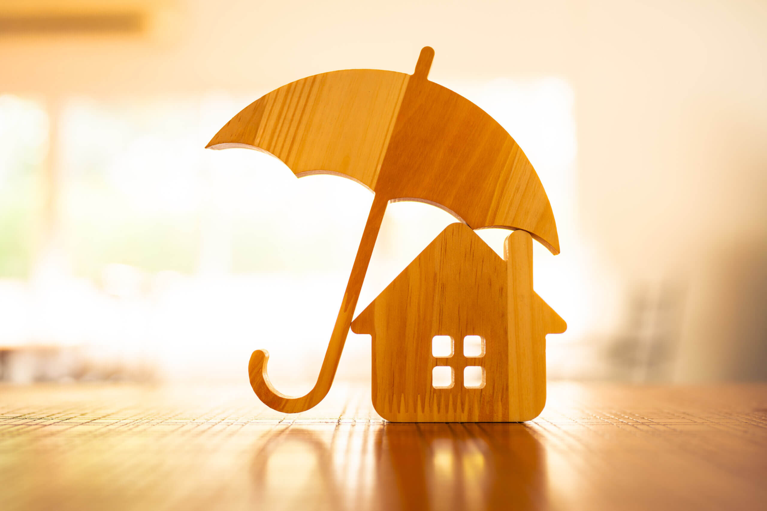 Wooden models depicting a house and an umbrella are placed on a wooden surface, with the umbrella positioned to shelter the house. The background is softly blurred, creating a warm, inviting ambiance.
