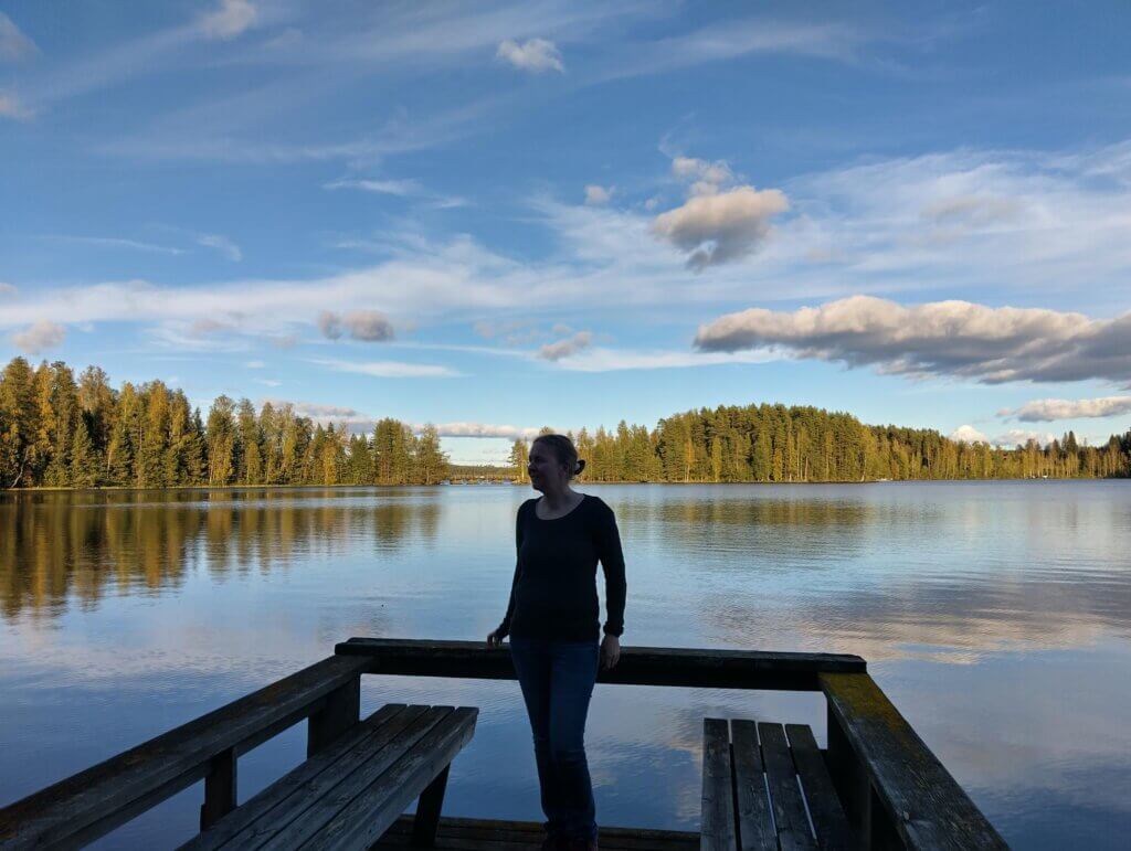 Nainen seisoo laiturilla järven äärellä, taustalla kauniit syksyiset maisemat ja kirkas taivas.