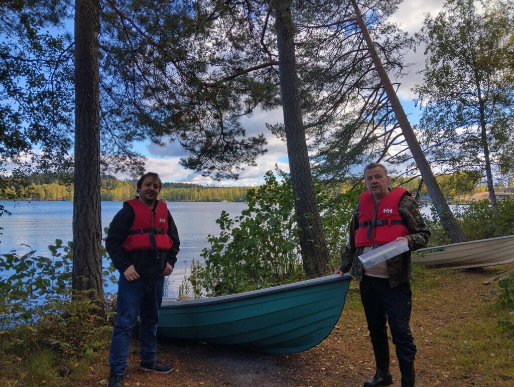 Kaksi miestä seisoo veneen vieressä järven rannalla, molemmilla on päällään punaiset pelastusliivit.