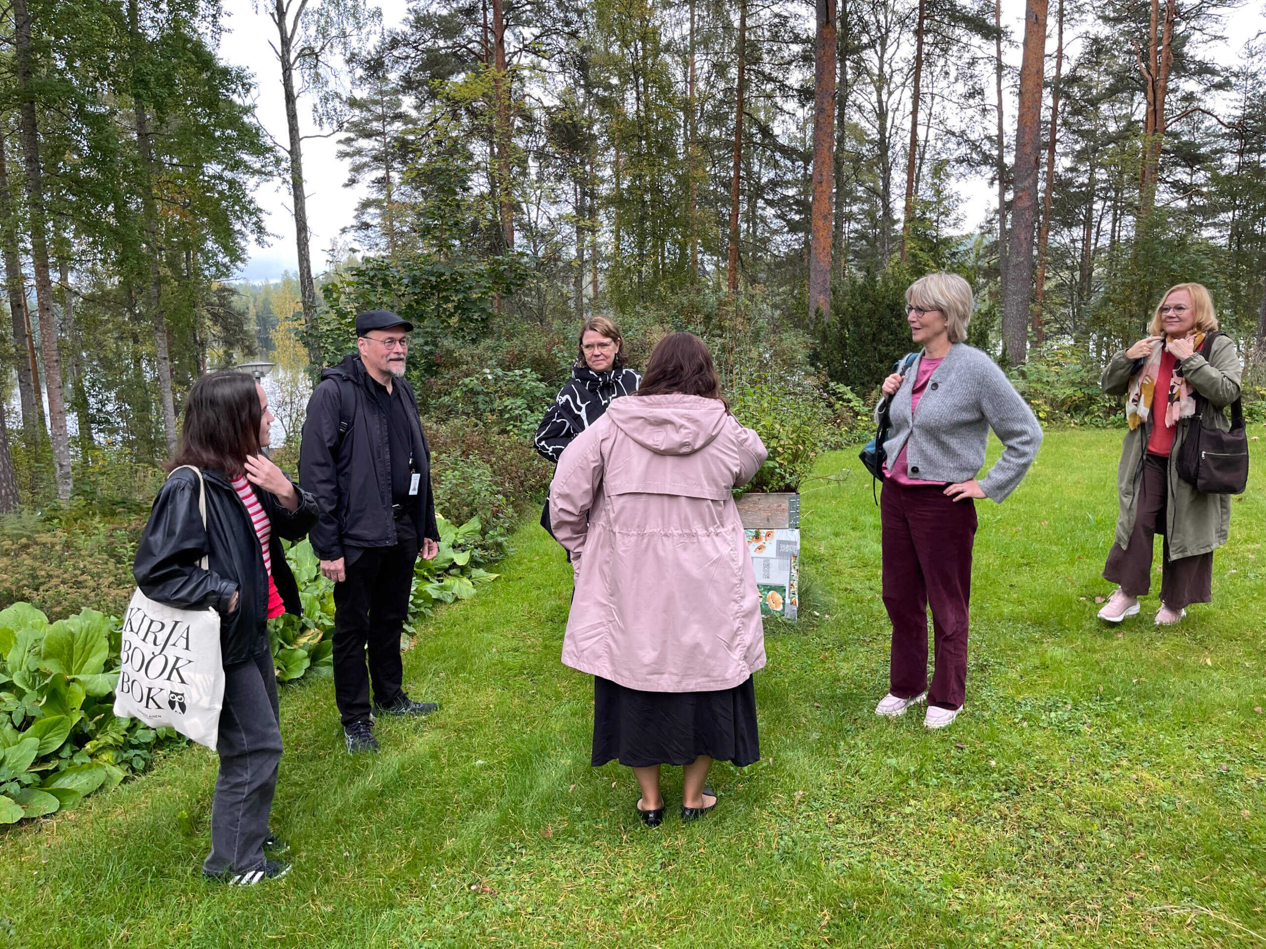 Ryhmä ihmisiä keskustelee ulkona metsäisessä ympäristössä järven rannalla.