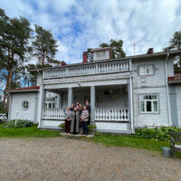 Ryhmä ihmisiä seisoo vanhan puutalon edessä, jossa on avara terassi ja ympärillä vihreää luontoa.
