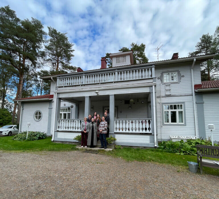 Ryhmä ihmisiä seisoo vanhan puutalon edessä, jossa on avara terassi ja ympärillä vihreää luontoa.