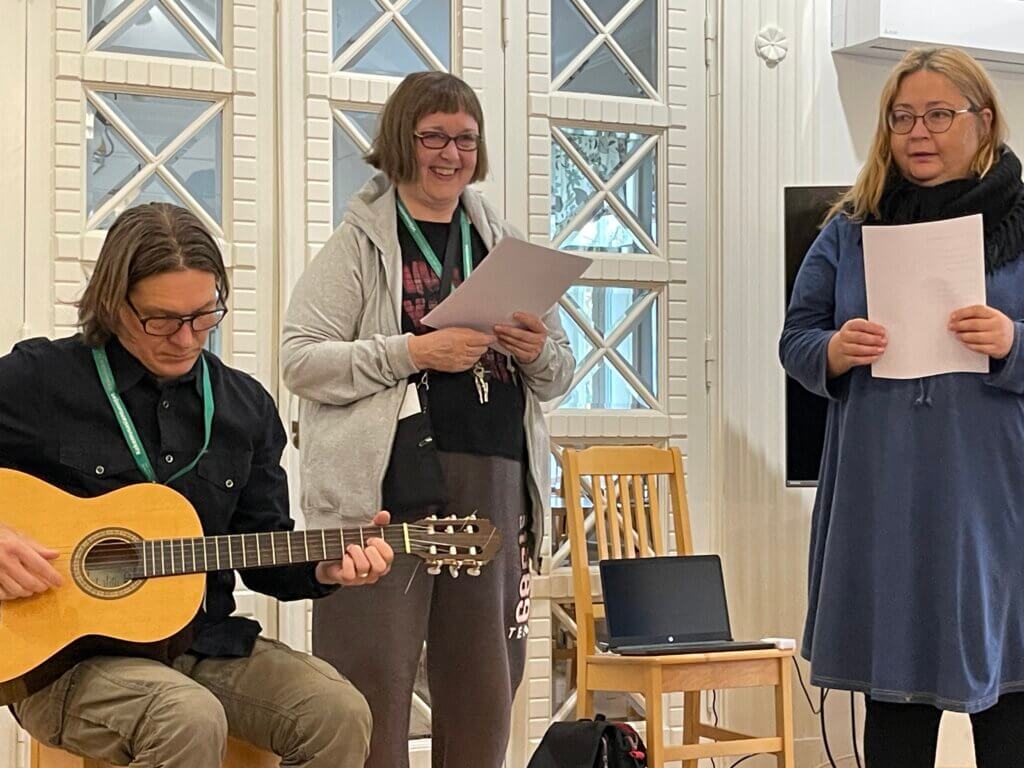 Kolme henkilöä esiintyy, yksi soittaa kitaraa ja kaksi muuta lukee paperista.