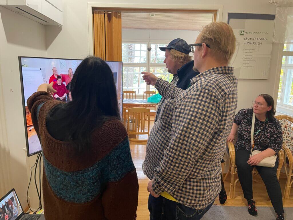 Neljä henkilöä katsoo televisiota ja osoittaa siinä näkyvää kuvaa, kun yksi henkilö istuu taustalla.