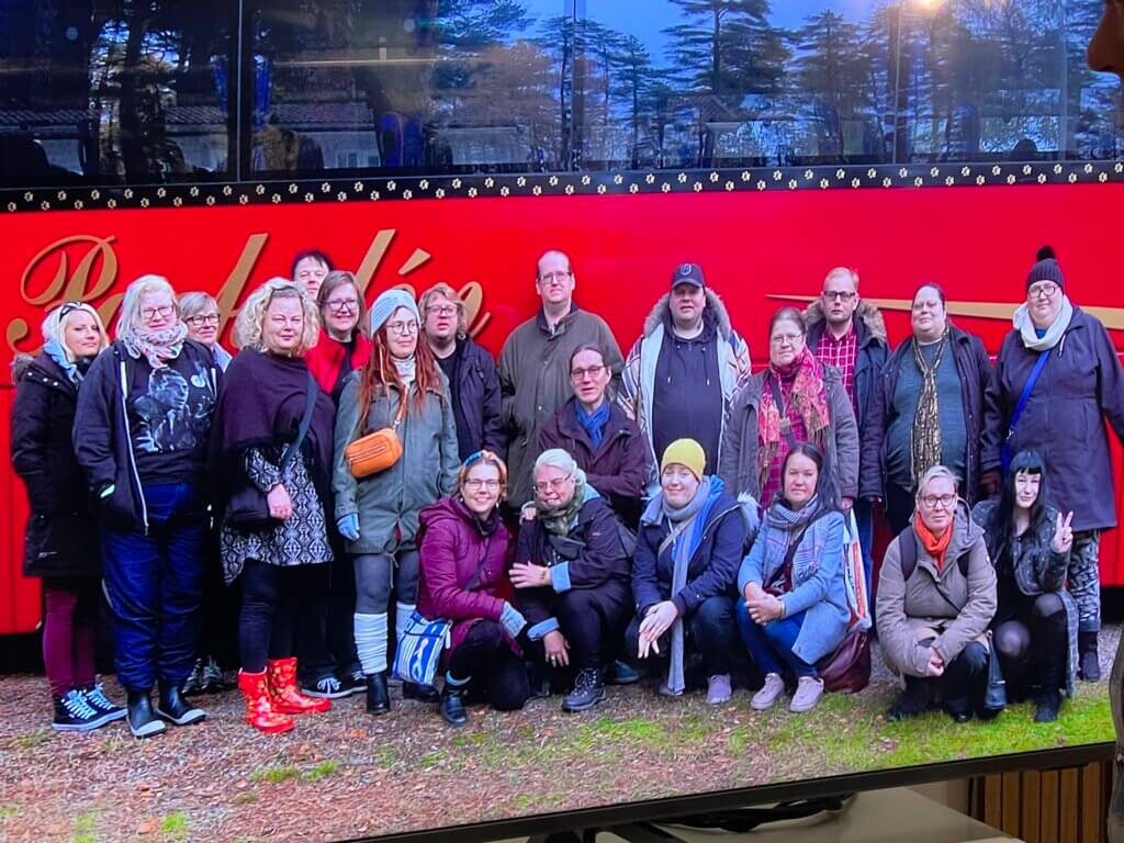 Ryhmä ihmisiä poseeraa punaisen bussin edessä metsäisessä ympäristössä.