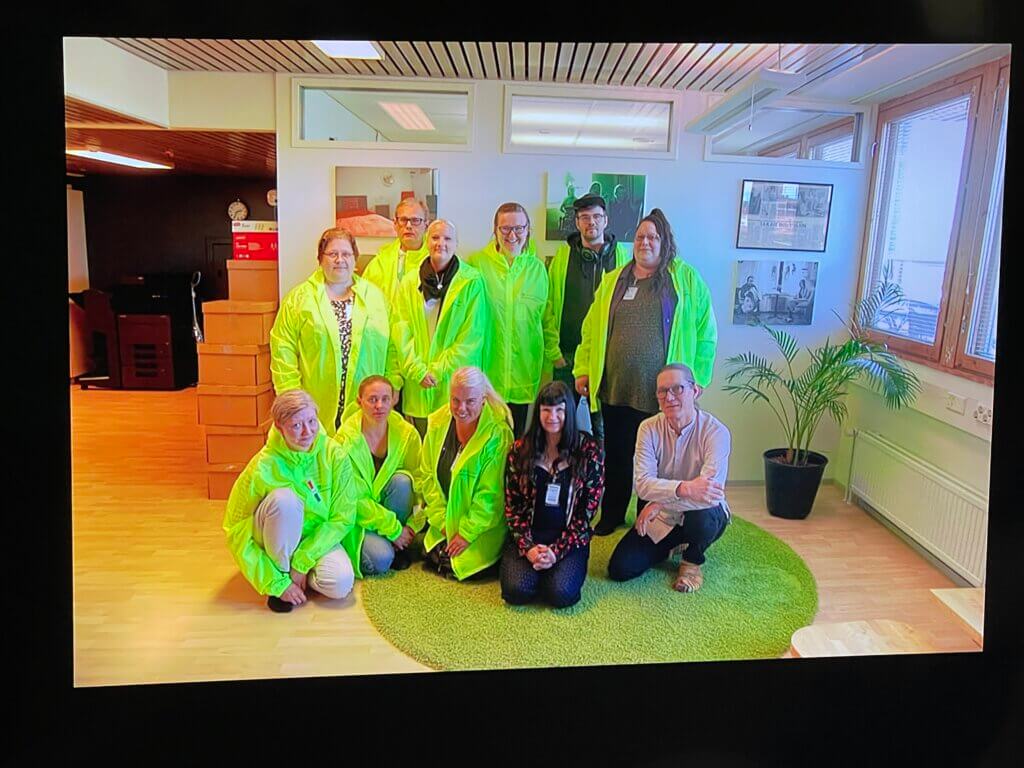 Kuvassa on ryhmä ihmisiä kirkkaan keltaisissa sadeviitoissa seisomassa ja istumassa vihreällä matolla toimistotilassa.