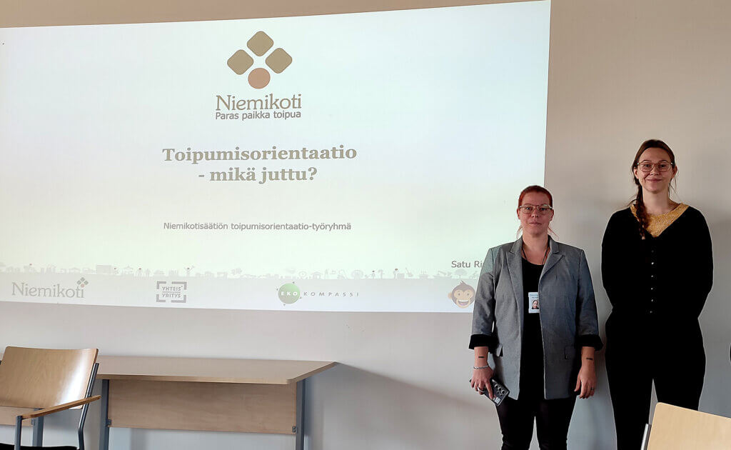 Kaksi naista seisoo esityksen edessä, jossa käsitellään toipumisorientaatiota Niemikotisäätiössä.
