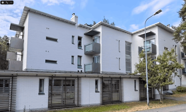 Moderni, vaaleaksi maalattu asuinrakennus, jossa on parvekkeita ja ympärillä vihreitä puita.
