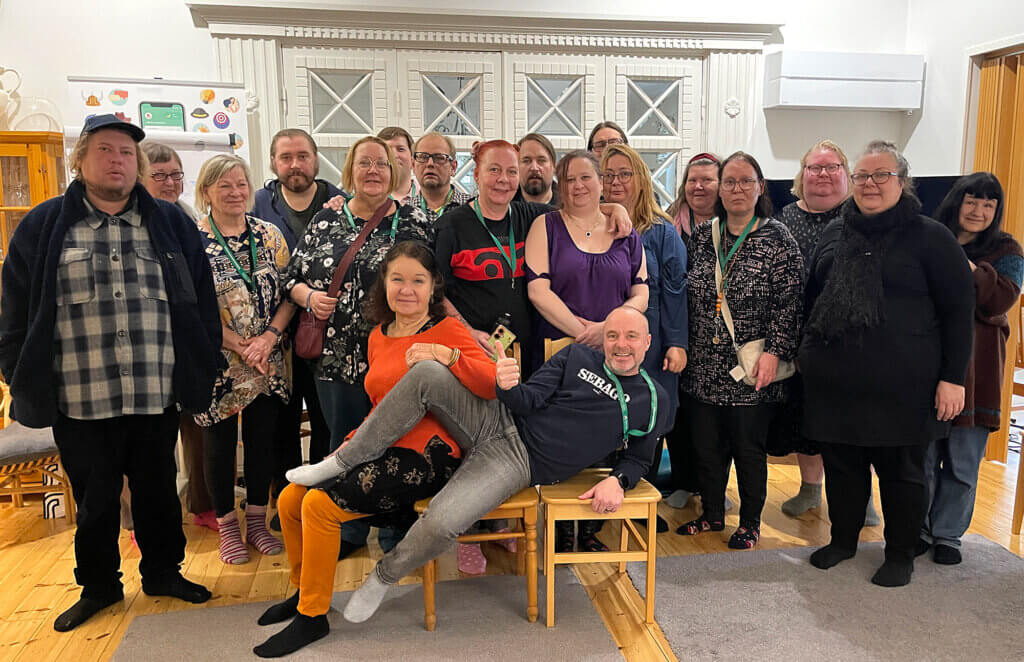 Ryhmä ihmisiä seisoo ja istuu yhdessä sisätilassa, monet heistä hymyilevät ja poseeraavat kameralle.