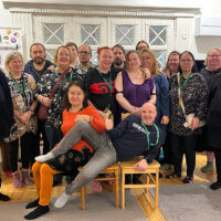 Ryhmä ihmisiä seisoo ja istuu yhdessä sisätilassa, monet heistä hymyilevät ja poseeraavat kameralle.