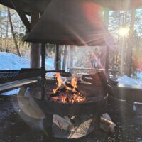 Nuotio palamassa metsäisessä ympäristössä, taustalla lumiset maisemat ja puut.