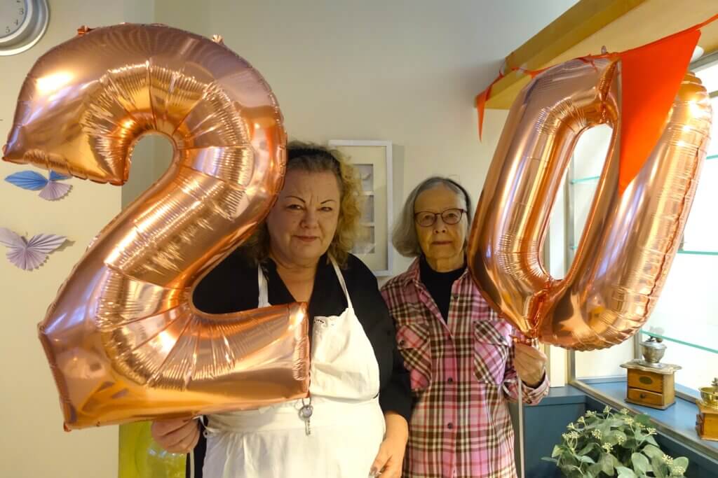 Two women hold large rose gold balloons shaped like the numbers 2 and 0, celebrating a special occasion in a decorated room.