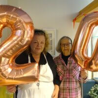 Two women hold large rose gold balloons shaped like the numbers 2 and 0, celebrating a special occasion in a decorated room.