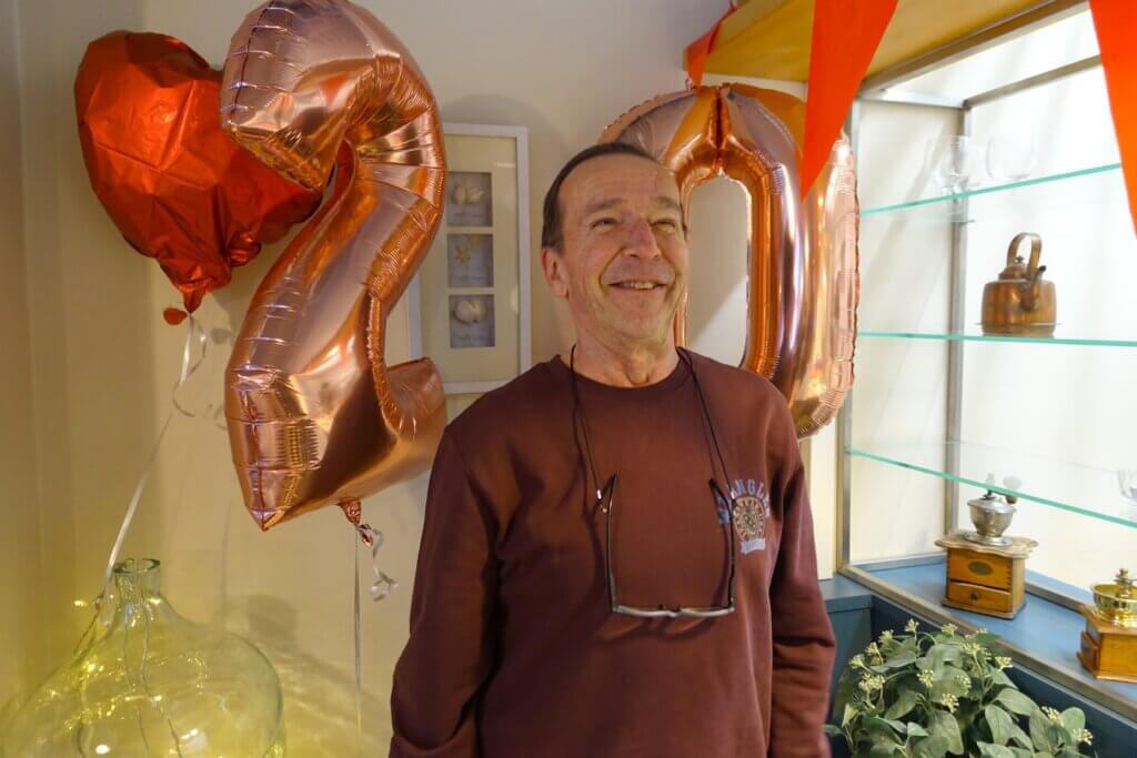 A smiling man stands between large rose gold balloons shaped like the numbers 2 and 0, with a heart-shaped balloon nearby.