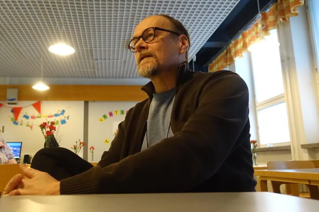 A man with glasses and a beard sits at a table in a brightly lit room decorated with flowers and colorful banners.