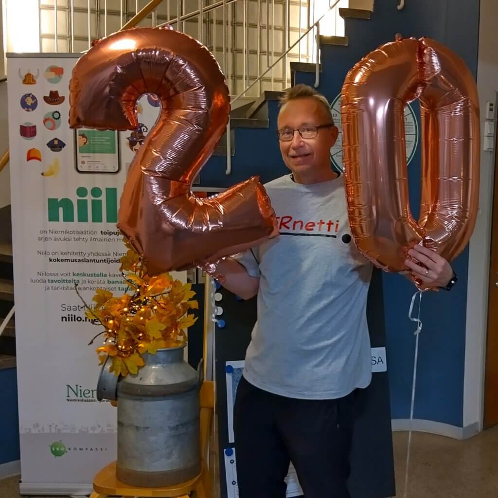 Mies pitää kädessään suuria, kuparinvärisiä ilmapalloja, joissa on numerot 2 ja 0, taustalla näkyy mainos ja koristeita.