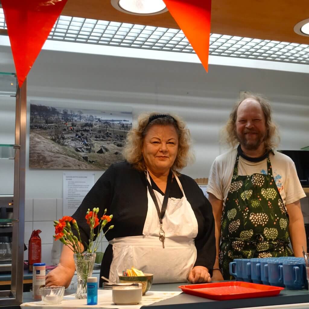 Two people, a woman in a black top and apron and a man in a patterned apron, stand behind a counter with food items and decorations, while a scenic photograph i