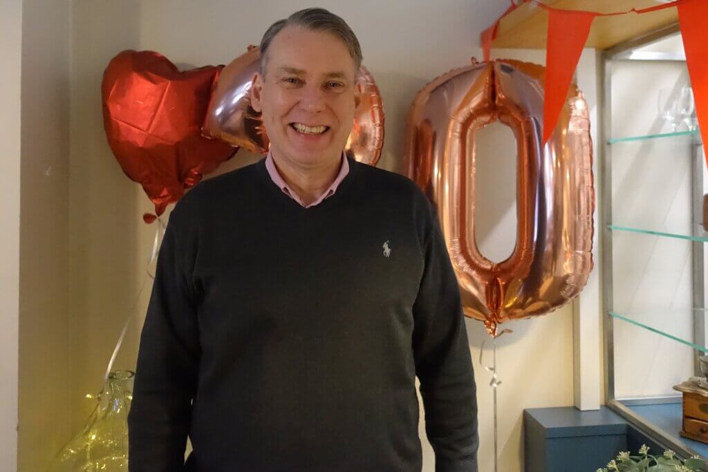 A smiling man in a dark sweater stands in front of large metallic balloons displaying the number "50" and red heart-shaped balloons.