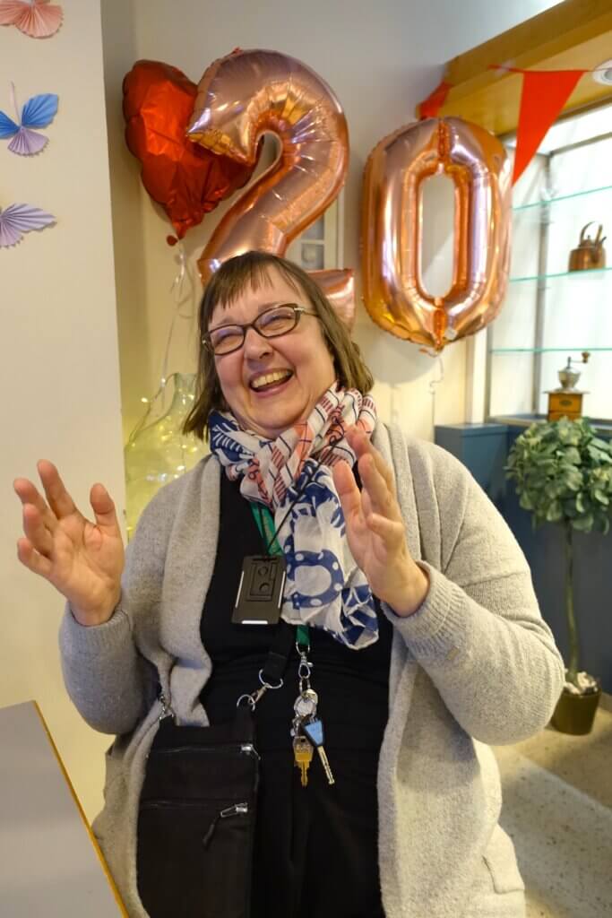 A smiling woman with glasses and a scarf gestures joyfully in front of large "20" balloons and decorative elements.