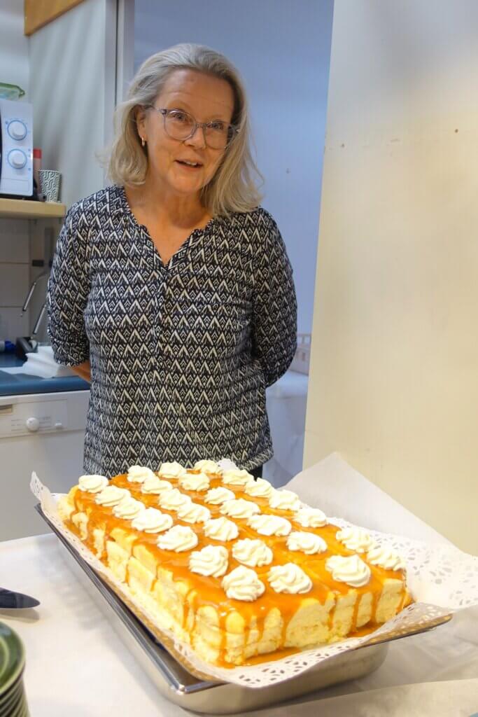 A woman with long, light hair and glasses smiles while standing behind a large cake topped with whipped cream and caramel sauce.