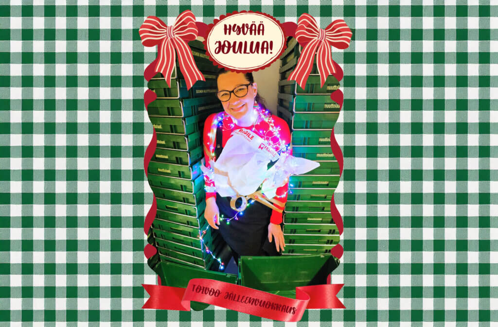 Nainen, joka on koristeltu jouluvaloilla ja pitää kädessään lahjakassia, istuu vihreiden laatikoiden keskellä, taustalla ruudullinen kuvio.