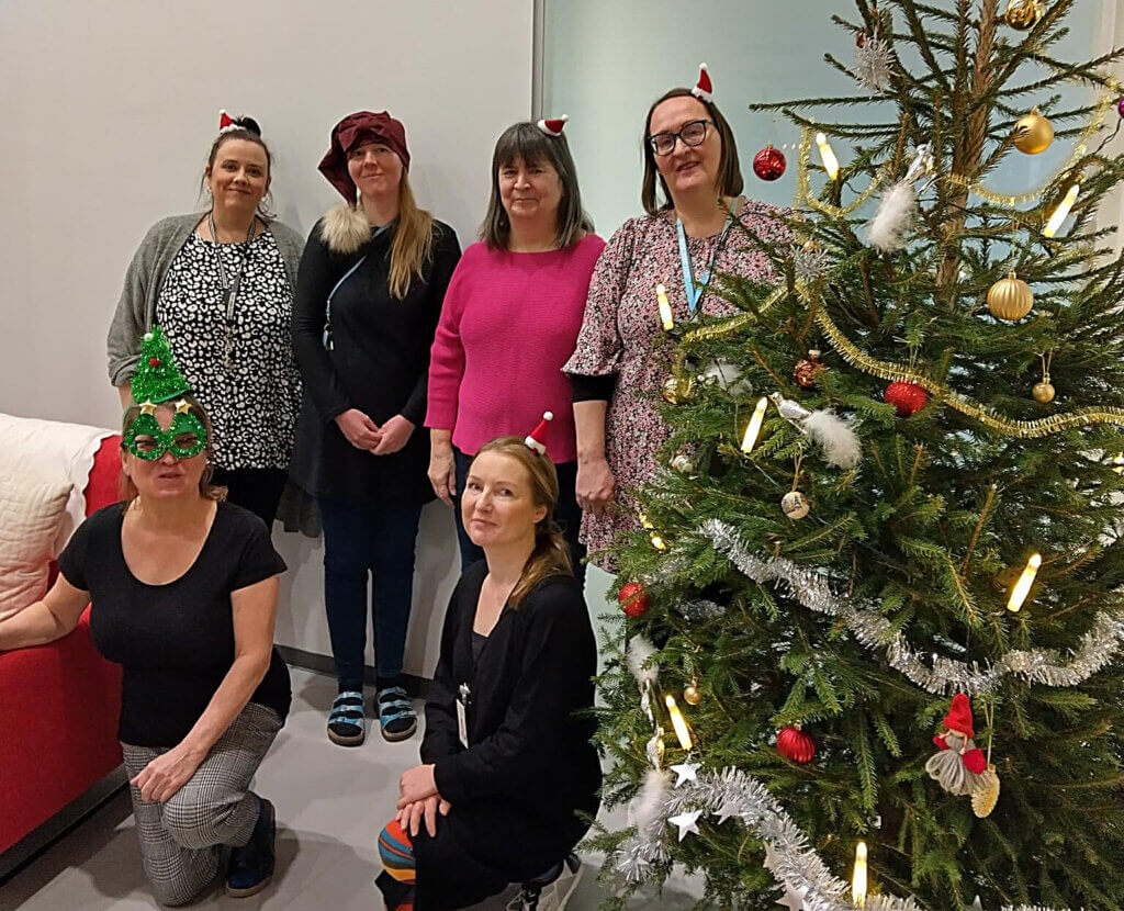 Kuudesta naisesta koostuva ryhmä, joka on pukeutunut jouluhattuihin, seisoo joulukuusen vieressä.