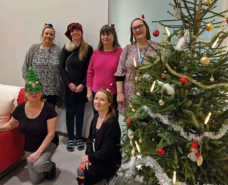 Kuudesta naisesta koostuva ryhmä, joka on pukeutunut jouluhattuihin, seisoo joulukuusen vieressä.