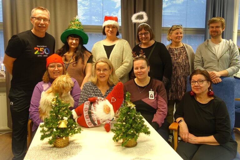 Ryhmä ihmisiä juhlii joulua koristeiden ja joulupuiden kanssa, mukana myös koira.