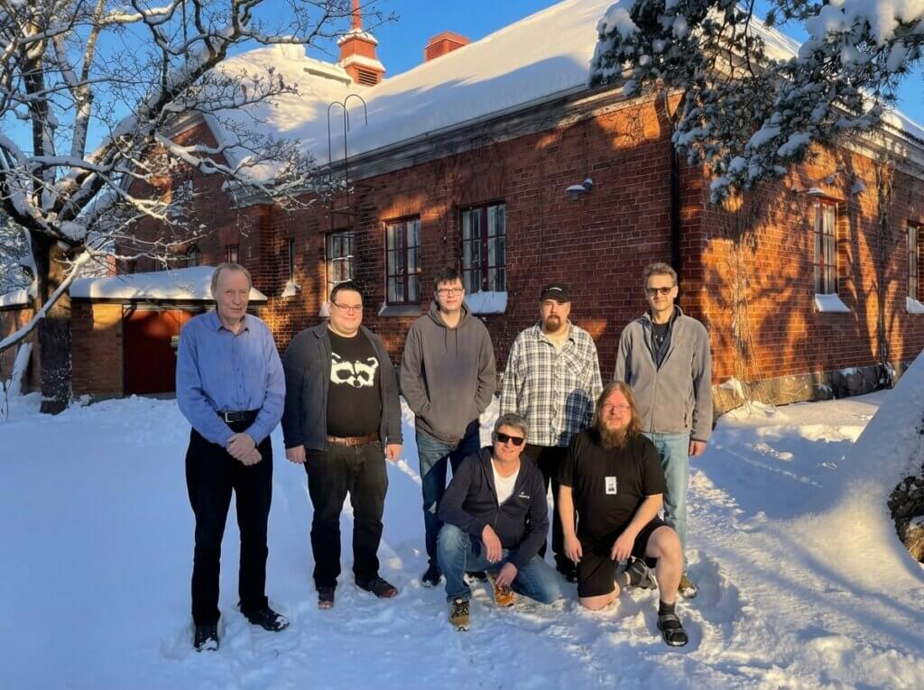 Ryhmän seitsemän henkilöä seisoo lumisessa maisemassa vanhan tiilirakennuksen edessä.