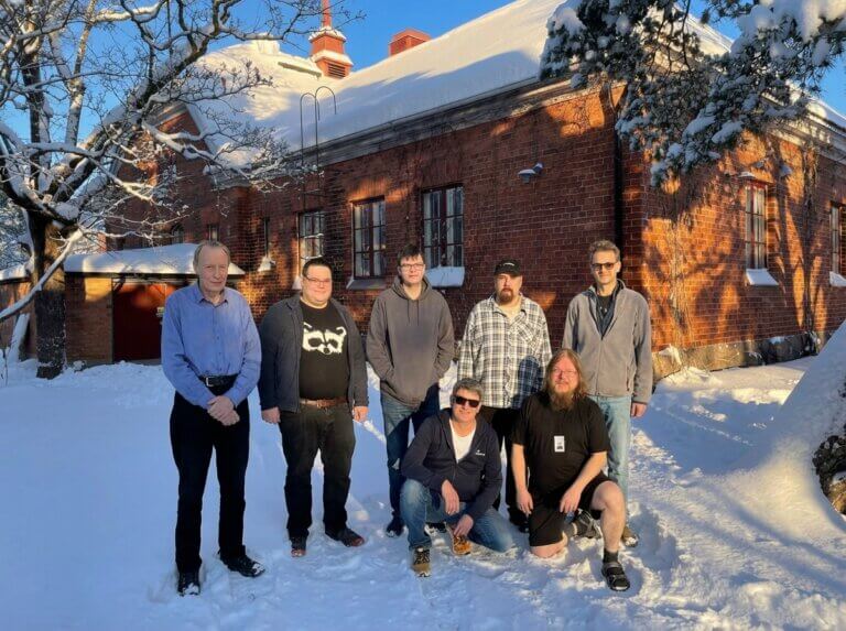 Ryhmän seitsemän henkilöä seisoo lumisessa maisemassa vanhan tiilirakennuksen edessä.
