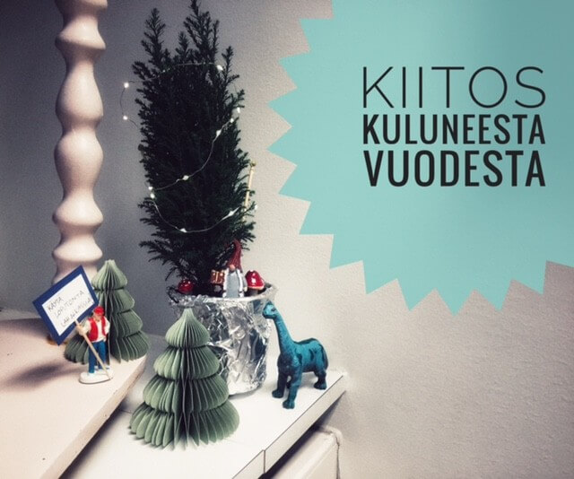 Pöydällä on koristeellinen joulukuusi, paperisia joulupuita ja sininen dinosaurus, sekä teksti "KIITOS KULUNEESTA VUODESTA".