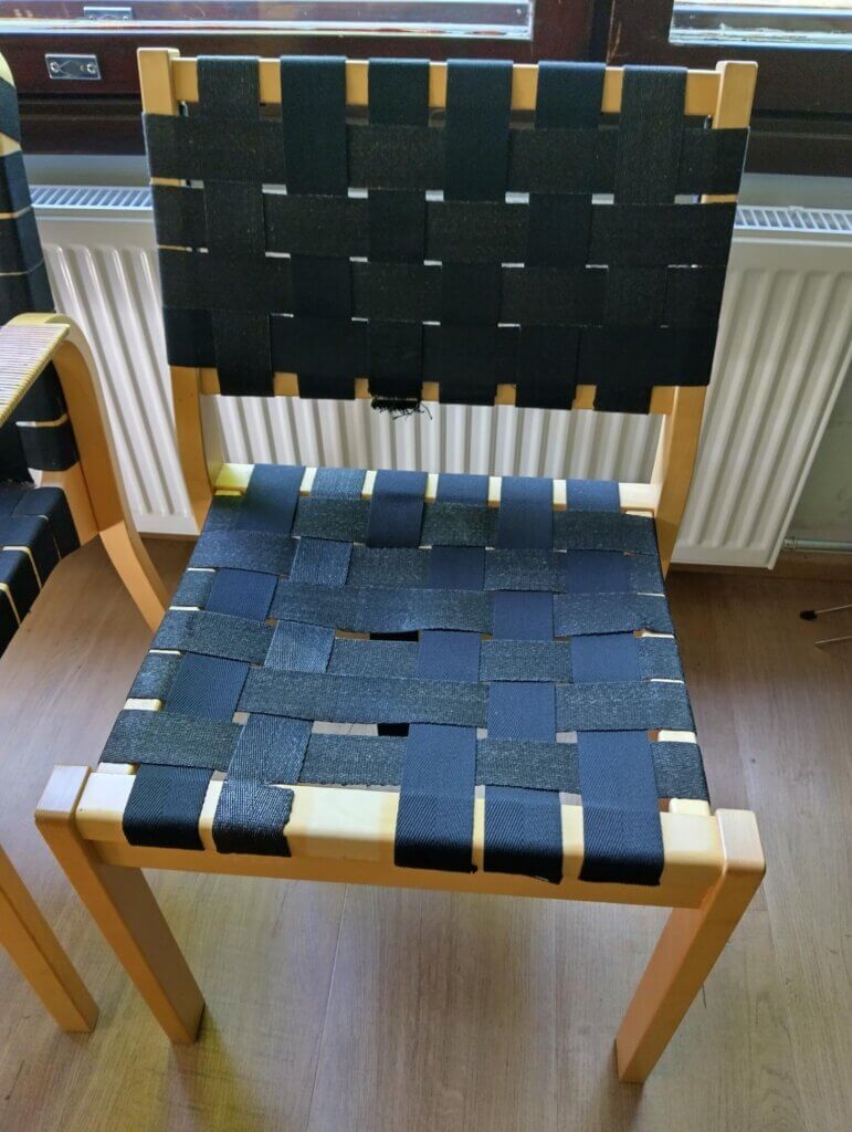 Tuoli, jossa on musta kudottu istuin ja selkänoja, sekä vaaleat puujalat.