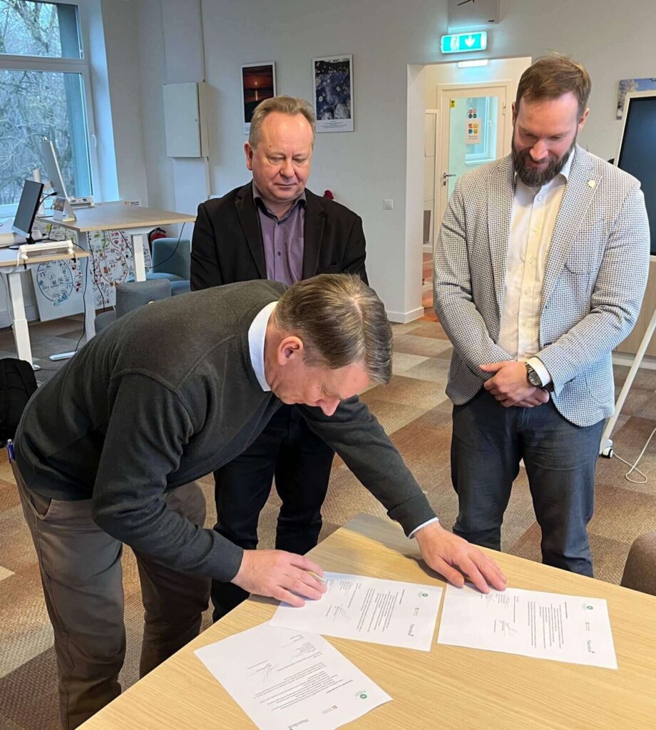 Kolme miestä allekirjoittaa asiakirjoja työtilassa, taustalla näkyy ikkunoita ja kalusteita.