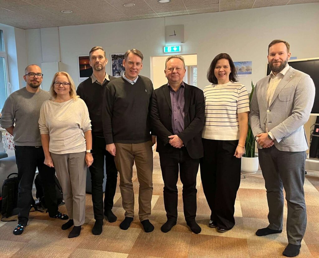 Ryhmä seitsemän henkilöä seisoo yhdessä sisätilassa, taustalla ikkuna ja esitystaulu.