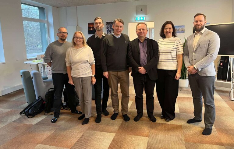 Ryhmä seitsemän henkilöä seisoo yhdessä sisätilassa, taustalla ikkuna ja esitystaulu.