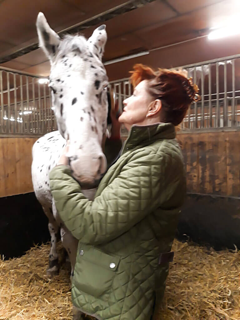 Nainen suutelee harmaata hevosta tallissa.