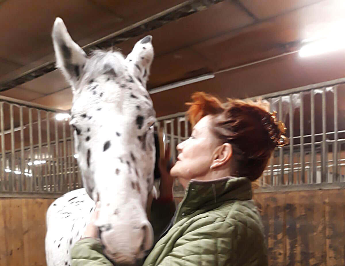 A woman with red hair gently interacts with a spotted horse in a stable.